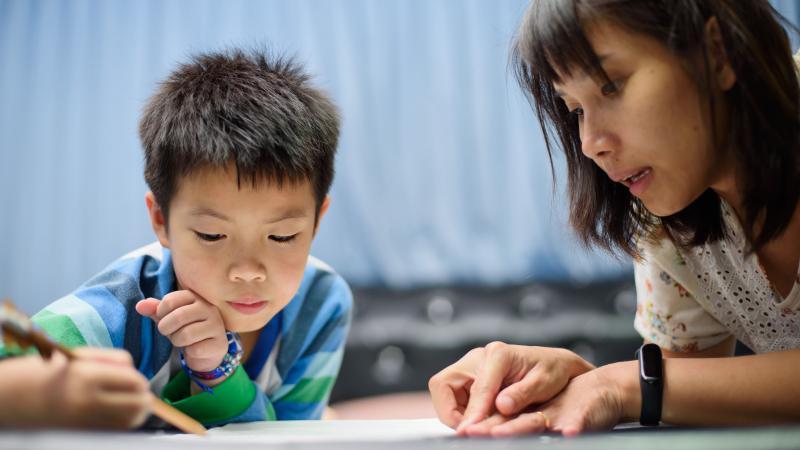 Child writes on paper. Woman points to something on that paper