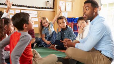 Classroom. Teacher and children are sitting on the floor. Teacher speaks to a boy.